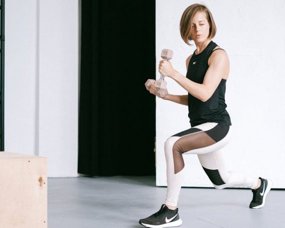 A young woman performing lunges with dumbbells in a modern indoor gym setting.